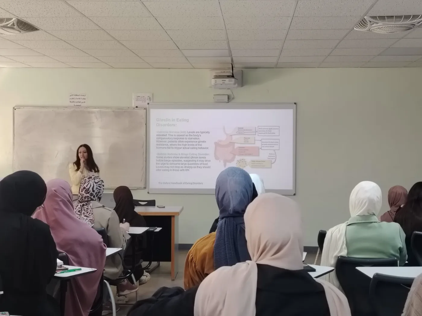 محاضرة أمام طلبة في قاعة دراسية مع عرض تقديمي